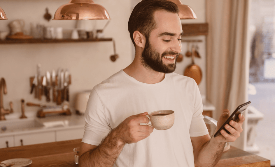 Smilende mann ser på telefon med kaffekopp i hånden